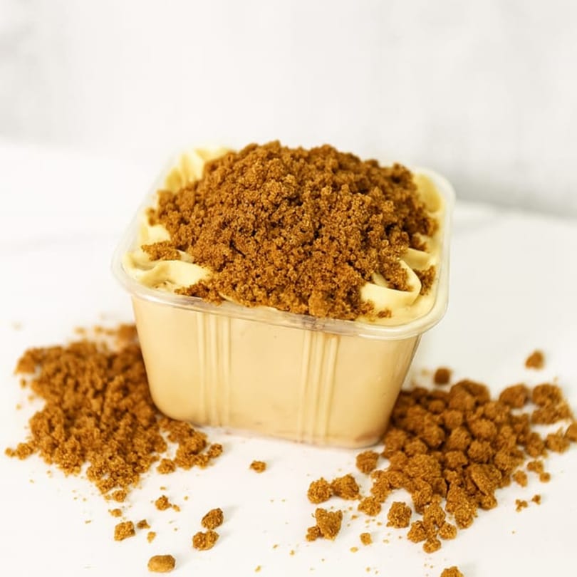 Sweet Kneads' square plastic container filled with light brown dessert and topped with cookie crumbles.