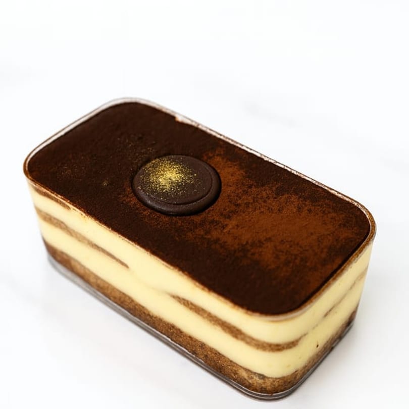 Sweet Kneads' tiramisu dessert in a clear rectangular container, topped with cocoa powder and a chocolate medallion.