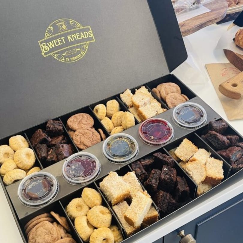 An open Sweet Kneads bakery box filled with cookies, brownies, cakes, and dipping sauces.