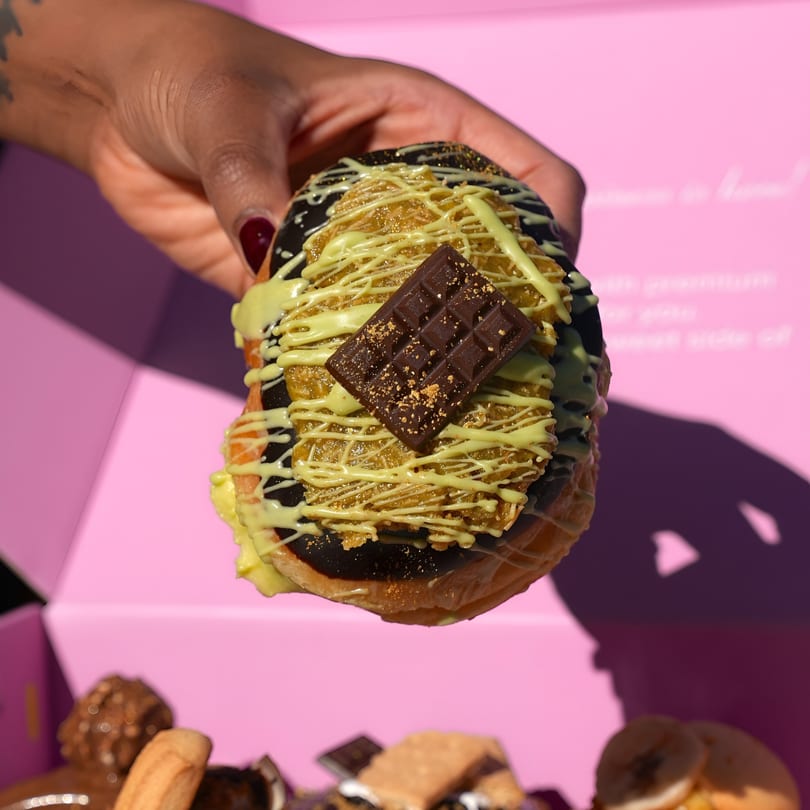 Hand holding a Sweet Kneads donut with dark chocolate, pistachio drizzle, and a chocolate bar.