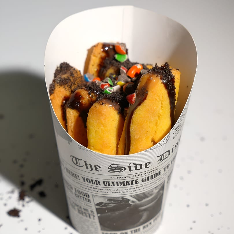 Sweet Kneads' churro-like pastries with chocolate drizzle, cookie crumbs, and colorful candies in a newspaper-print cone.