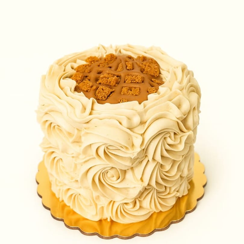 A small, round cake with swirled beige frosting, topped with caramel and cookie crumbles.