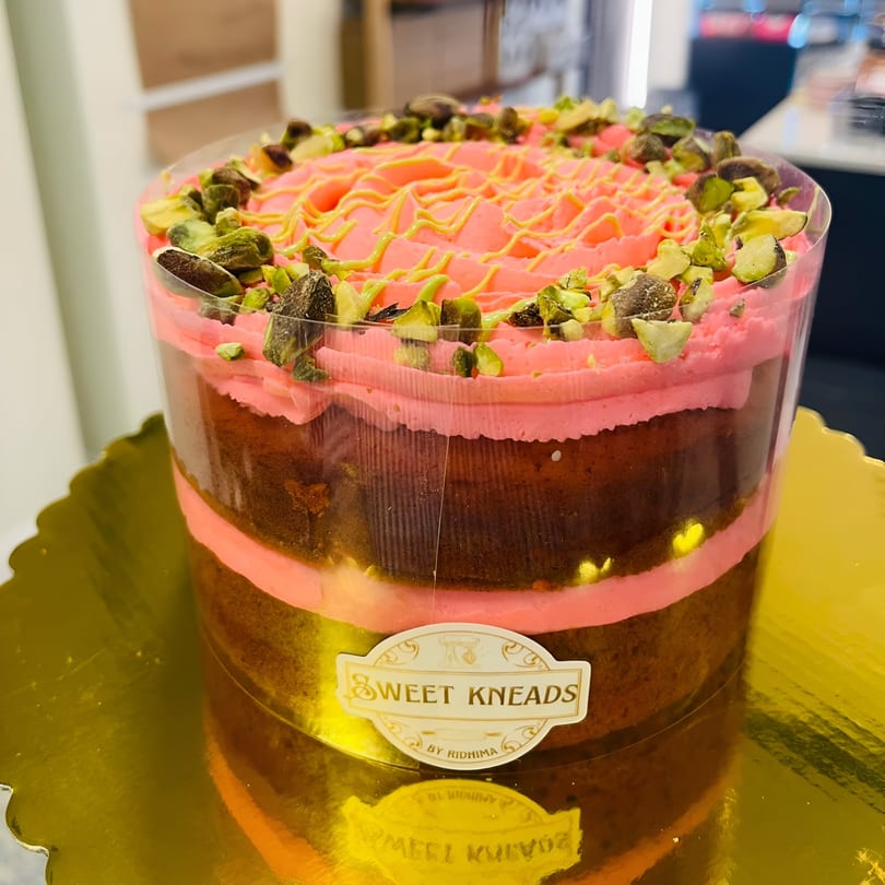 Sweet Kneads' layered cake with pink frosting, yellow drizzle, and pistachio crumbles on a gold platter.